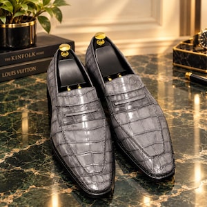 Peut inclure: Une paire de mocassins gris avec un motif texturé en peau de crocodile. Chaque chaussure présente une bride de mocassin classique sur l'empeigne, un bout pointu et une doublure intérieure noire avec un insert métallique doré au talon.