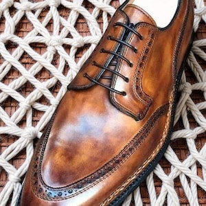 May include: A brown leather dress shoe with intricate stitching and a classic lace-up design. The shoe features a polished finish and a decorative toe cap. A silver ring rests inside the shoe. The shoe is displayed on a woven surface.