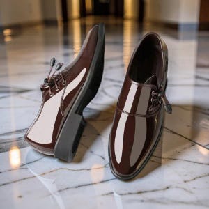Può includere: Un paio di scarpe eleganti in pelle marrone con finitura lucida. Le scarpe hanno un classico design stringato e un aspetto elegante e lucido. Le suole sono grigio scuro e le scarpe sono appoggiate su una superficie di marmo.