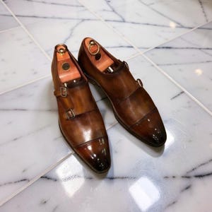 Puede incluir: Un par de zapatos marrones de cuero con doble hebilla y hormas de madera en el interior. Los zapatos tienen un acabado brillante y hebillas plateadas. Los zapatos están colocados sobre una superficie de baldosas de mármol blanco.
