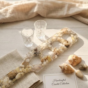 May include: A clear resin serving paddle filled with seashells and small stones, with three circular cutouts. Two small clear glasses and a card reading "Handcrafted Coastal Collection" are present. Several seashells and a linen napkin are nearby.