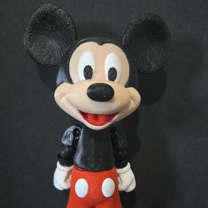 May include: A 3D-printed Mickey Mouse figurine. The figure has large black ears, a white face, and a black nose. It wears a black top, red shorts with white buttons, white gloves, and yellow shoes. The figurine is standing.