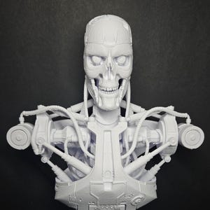 May include: A white, detailed bust of a skeletal robot with exposed mechanical components. The sculpture features a skull-like head, visible teeth, and intricate wiring and machinery on the chest and shoulders. The bust is mounted on a geometric base.