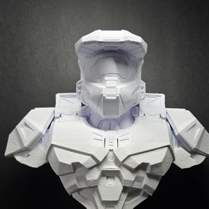 Puede incluir: Busto impreso en 3D de una figura futurista blindada, predominantemente blanco. La escultura detallada presenta un casco, una armadura de pecho y hombreras, con texturas y diseños geométricos visibles. Una representación detallada.
