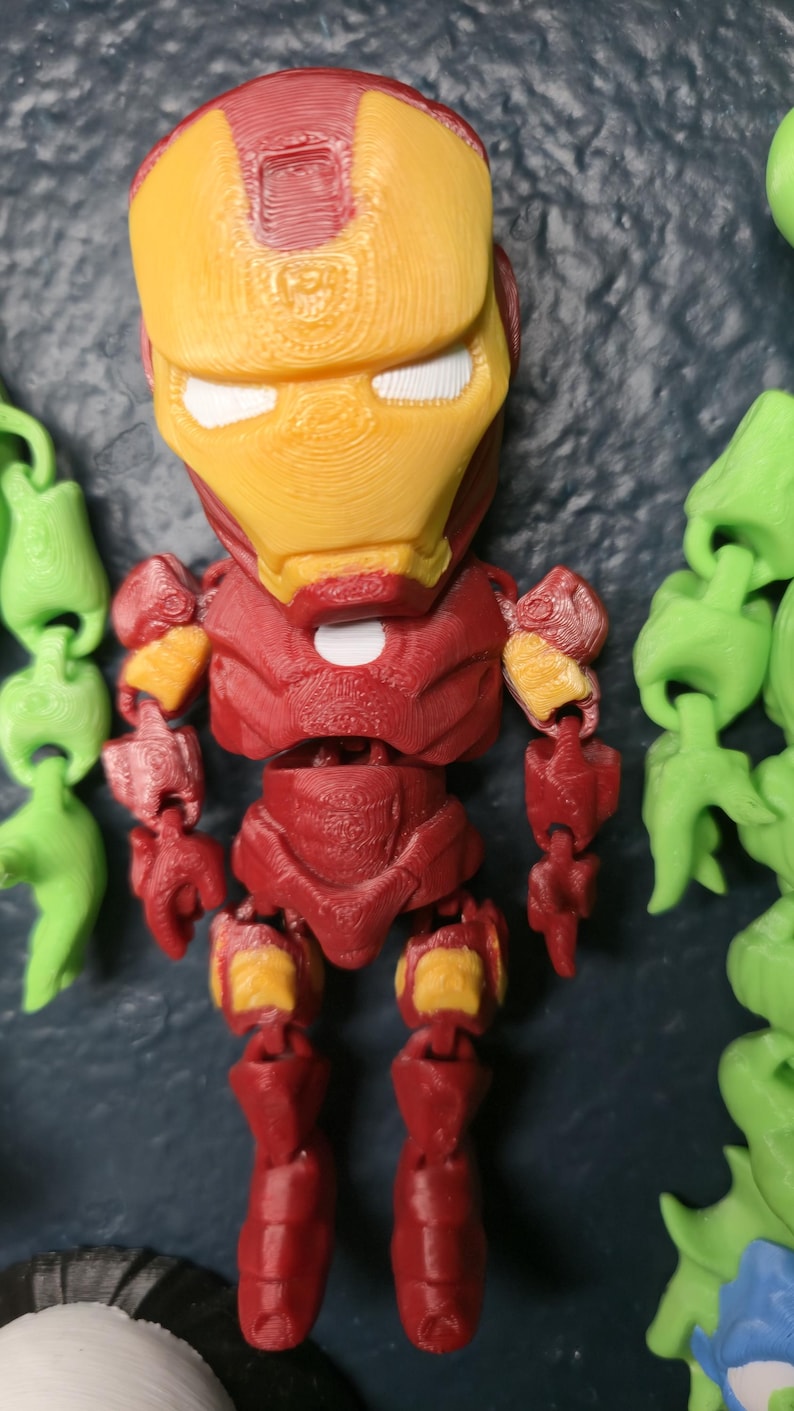 May include: A 3D-printed Iron Man figurine in a standing pose. The figure is primarily red with yellow accents on the helmet and joints. The eyes are white. The figure is a stylized, cartoon-like representation of the superhero.