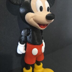 May include: A 3D-printed Mickey Mouse figurine. The figure features a black body, ears, and nose, with a tan face and white gloves. It wears red shorts with white buttons and yellow shoes. The figurine is a classic Disney character.