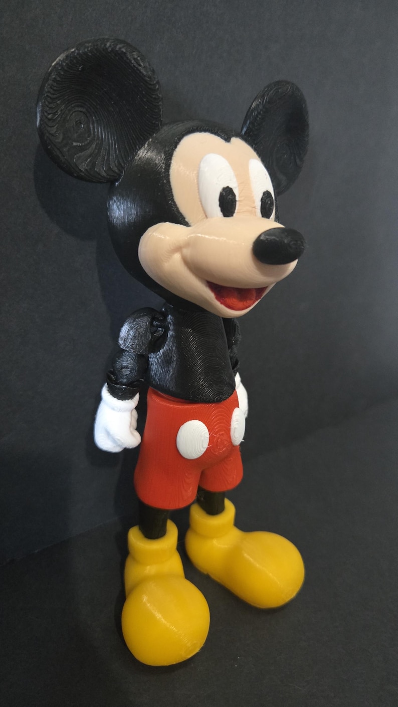 May include: A 3D-printed Mickey Mouse figurine. The figure features a black body, ears, and nose, with a tan face and white gloves. It wears red shorts with white buttons and yellow shoes. The figurine is a classic Disney character.