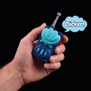 Pode incluir: Um dispositivo de controlo remoto retangular azul-escuro com um emblema de concha azul-claro na parte frontal, uma antena cinzenta e três pequenos botões azul-claros. É visível um balão de fala azul-claro com o texto Clicker!