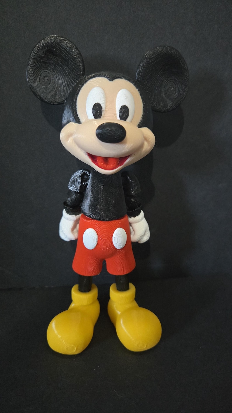 May include: A 3D-printed Mickey Mouse figurine. The figure has large black ears, a white face, and a black nose. It wears a black top, red shorts with white buttons, white gloves, and yellow shoes. The figurine is standing.