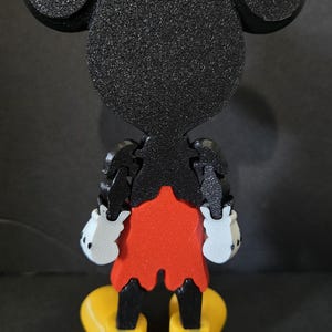 May include: A 3D-printed Mickey Mouse figure, viewed from the back. The figure features black ears, head, and arms, a red shorts, white gloves, and yellow shoes. The figure is made of textured plastic.