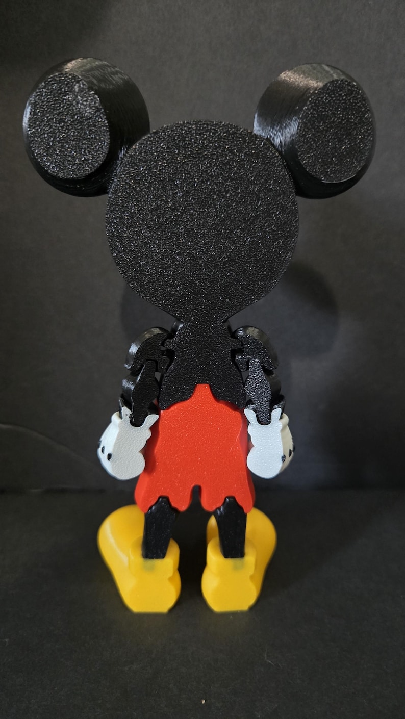 May include: A 3D-printed Mickey Mouse figure, viewed from the back. The figure features black ears, head, and arms, a red shorts, white gloves, and yellow shoes. The figure is made of textured plastic.