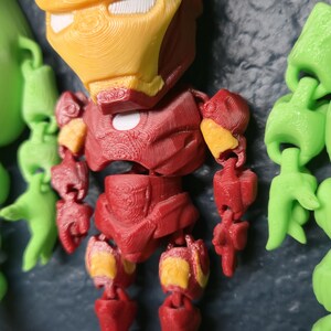 May include: A 3D-printed Iron Man figure, featuring a red and yellow color scheme. The figure is articulated and has a detailed design, showcasing the iconic superhero's helmet and armor. The figure is part of a collection of other figures.