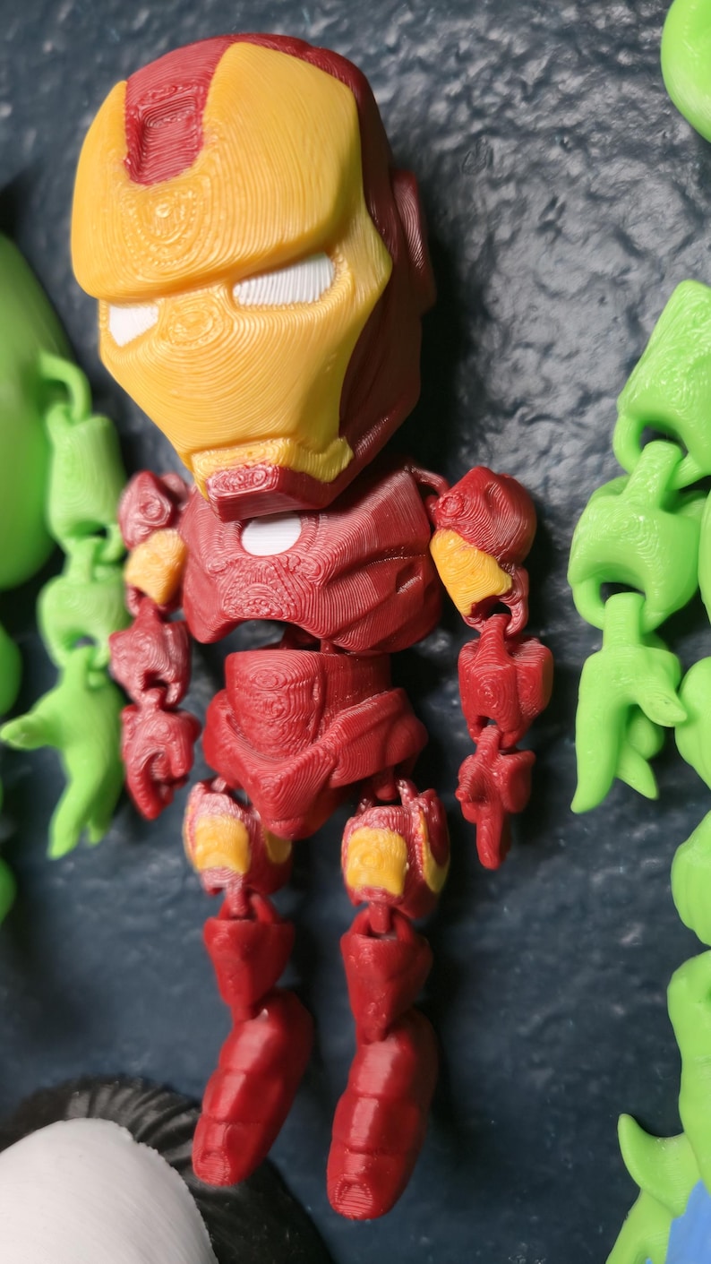May include: A 3D-printed Iron Man figure, featuring a red and yellow color scheme. The figure is articulated and has a detailed design, showcasing the iconic superhero's helmet and armor. The figure is part of a collection of other figures.
