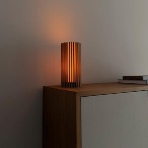 Op de afbeelding: Een cilindervormige tafellamp opgebouwd uit verticale houten latten in een cirkelvormig patroon. De lamp verspreidt een warme, zachte oranje gloed van binnenuit, waardoor de ruimte tussen de latten wordt verlicht en licht op de muur valt.