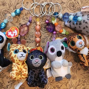 May include: A collection of plush animal keychains, including a panda, giraffe, leopard, koala, tiger, and a seal. Each keychain has a beaded strand and a metal ring. The animals are colorful and have large, glittery eyes.