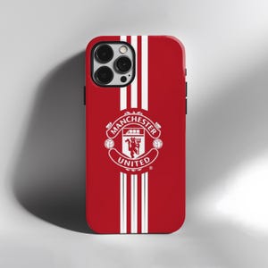 May include: Red phone case with white vertical stripes and the Manchester United logo. The logo is in white and features the team's crest and the words "Manchester United". The case has a glossy finish.