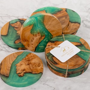 May include: A set of wooden coasters with a teal resin inlay. The coasters are round, with natural wood grain and a smooth, glossy finish. A tag with the text "HIGHLAND HOME RESIN" is attached to a stack of coasters.