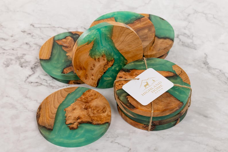 May include: A set of wooden coasters with a teal resin inlay. The coasters are round, with natural wood grain and a smooth, glossy finish. A tag with the text "HIGHLAND HOME RESIN" is attached to a stack of coasters.