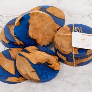 May include: Set of round wooden coasters with a blue resin inlay. The coasters have a natural wood grain pattern and are bundled together with twine. A tag with the words "Highland Home" is attached.