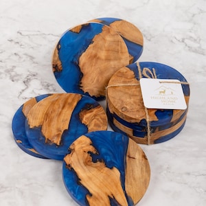 May include: A collection of round coasters with a unique design. Each coaster features a combination of natural wood and vibrant blue resin. The coasters are bundled together with twine and a tag that reads "HIGHLAND HOME RESIN".