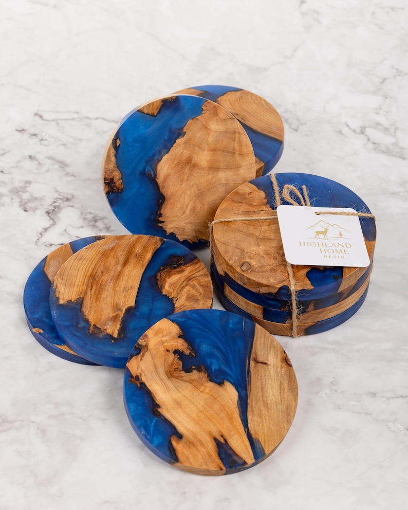 May include: A collection of round coasters with a unique design. Each coaster features a combination of natural wood and vibrant blue resin. The coasters are bundled together with twine and a tag that reads "HIGHLAND HOME RESIN".