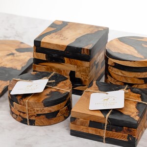 May include: Wooden coasters and a cutting board with a unique design. The coasters are square, round, and hexagonal, featuring a combination of natural wood and black resin. Each coaster set is tied with twine and has a tag.