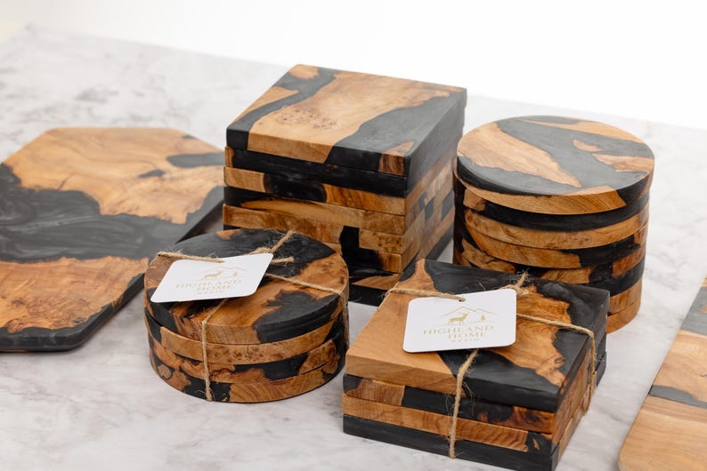 May include: Wooden coasters and a cutting board with a unique design. The coasters are square, round, and hexagonal, featuring a combination of natural wood and black resin. Each coaster set is tied with twine and has a tag.