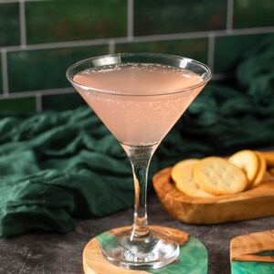 May include: A pink cocktail in a stemmed glass sits on a wooden and teal resin coaster. The drink is in a classic martini glass. Crackers are on a wooden serving board in the background. The background includes a green tile wall and a dark green cloth.