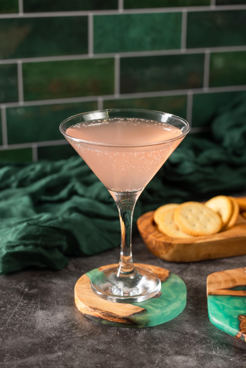 May include: A pink cocktail in a stemmed glass sits on a wooden and teal resin coaster. The drink is in a classic martini glass. Crackers are on a wooden serving board in the background. The background includes a green tile wall and a dark green cloth.