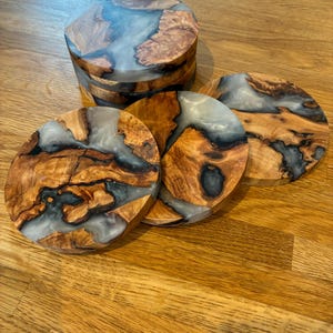 May include: A set of handcrafted wooden coasters with a unique design. The coasters feature a combination of natural wood grain and a clear, resin-like material. The wood has a warm, brown tone, while the resin appears translucent with a hint of blue. The coasters are round.