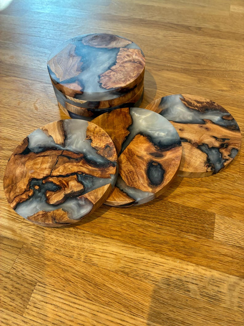 May include: A set of handcrafted wooden coasters with a unique design. The coasters feature a combination of natural wood grain and a clear, resin-like material. The wood has a warm, brown tone, while the resin appears translucent with a hint of blue. The coasters are round.
