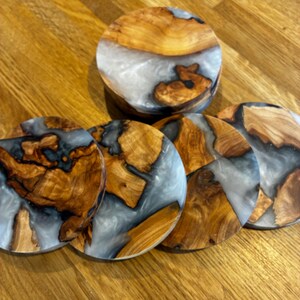May include: A set of six wooden coasters with a unique design. The coasters feature a combination of light brown wood, gray resin, and a circular shape. One coaster is stacked on top of the others. The coasters are on a wooden surface.