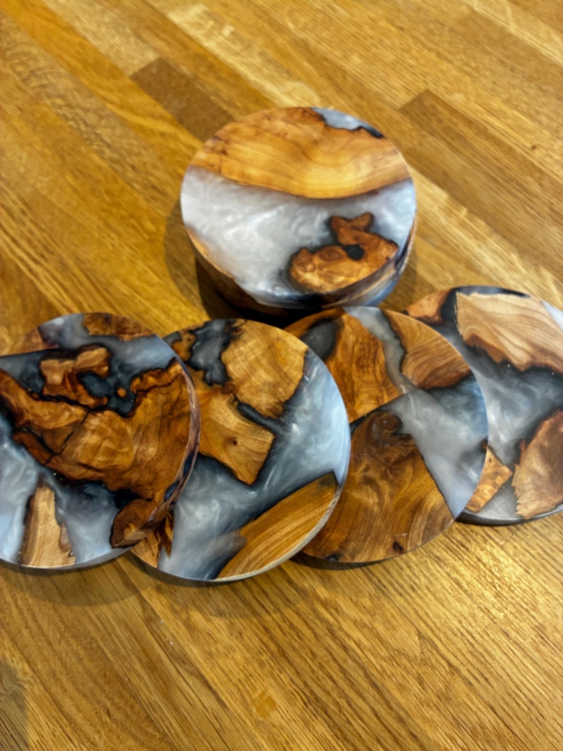 May include: A set of six wooden coasters with a unique design. The coasters feature a combination of light brown wood, gray resin, and a circular shape. One coaster is stacked on top of the others. The coasters are on a wooden surface.