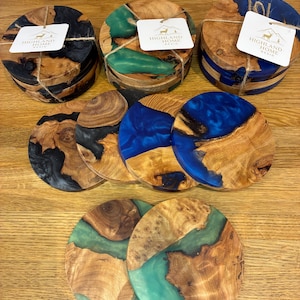 May include: Wooden coasters with epoxy resin in various colors, including black, green, and blue. Each coaster is round and features a unique wood grain pattern. Some sets are tied with twine and include a tag that reads "HIGHLAND HOME RESIN".