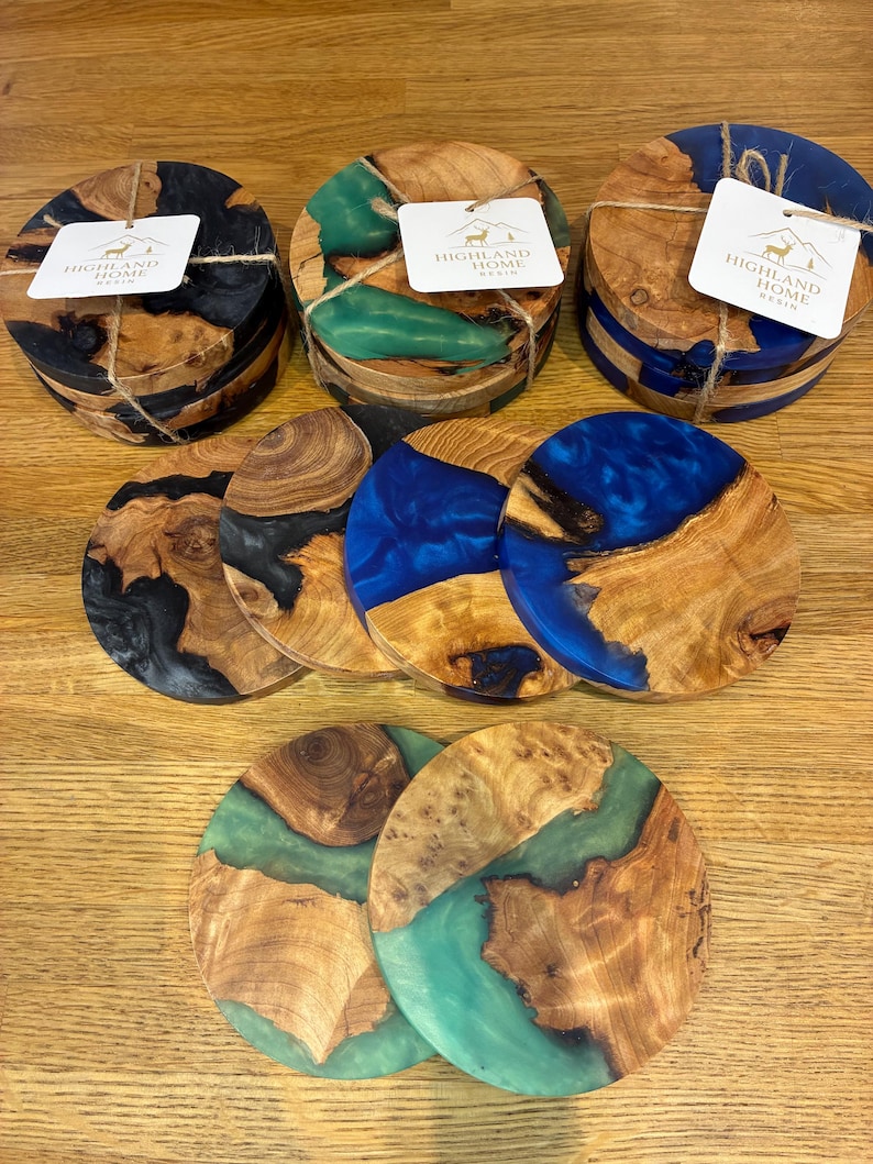 May include: Wooden coasters with epoxy resin in various colors, including black, green, and blue. Each coaster is round and features a unique wood grain pattern. Some sets are tied with twine and include a tag that reads "HIGHLAND HOME RESIN".