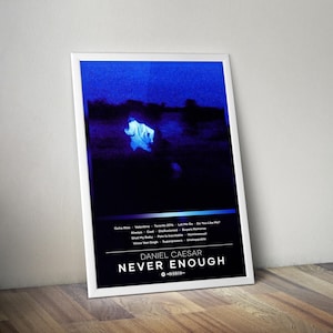 May include: A framed poster with a blue and black image of a figure in a white garment. The poster has the text "DANIEL CAESAR NEVER ENOUGH" in white. Below the image are song titles. The poster is leaning against a wooden surface.