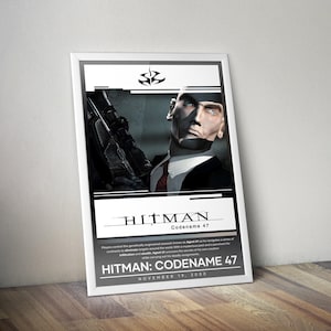 May include: A framed poster for the video game "Hitman: Codename 47." The poster features a bald man in a suit and tie holding a gun. The title "HITMAN" is prominently displayed, along with the release date, November 19, 2000.