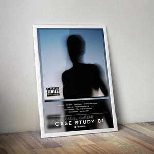 May include: A framed poster featuring a blurred silhouette of a person against a blue and white gradient background. The poster includes the text "PARENTAL ADVISORY EXPLICIT CONTENT" and song titles. The artist's name "DANIEL CAESAR" and album title "CASE STUDY 01" are also displayed.