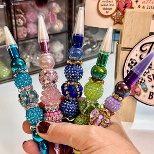 Könnte beinhalten: Fünf Stifte mit dekorativen Perlengehäusen in verschiedenen Texturen und Farben, darunter Türkis, Pink, Blau, Grün und Lila. Jeder Stift hat eine transparente Kunststoffkappe und ist mit metallischen Zwischenstücken sowie facettierten Perlen verziert.