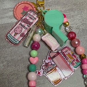 Puede incluir: Un llavero con cierre dorado, un silbato verde menta y varios dijes que incluyen una bebida energ&eacute;tica Alani Nu rosa, un dulce rosa con la etiqueta Sweeet y un dije de vaso con purpurina, todo decorado con cuentas rosas, verdes y transparentes.