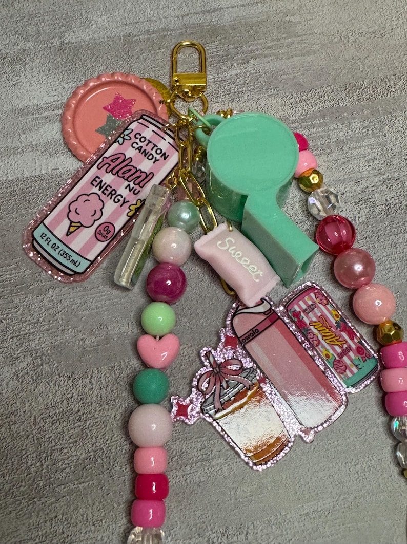 Puede incluir: Un llavero con cierre dorado, un silbato verde menta y varios dijes que incluyen una bebida energ&eacute;tica Alani Nu rosa, un dulce rosa con la etiqueta Sweeet y un dije de vaso con purpurina, todo decorado con cuentas rosas, verdes y transparentes.