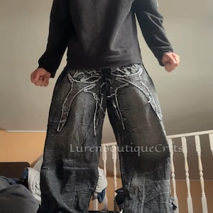 May include: Black sweatshirt with a silver emblem and distressed black denim jeans. The jeans have white stitching and a wide leg design. White trainers complete the outfit. The text "LurenBoutiqueCrfts" is visible.