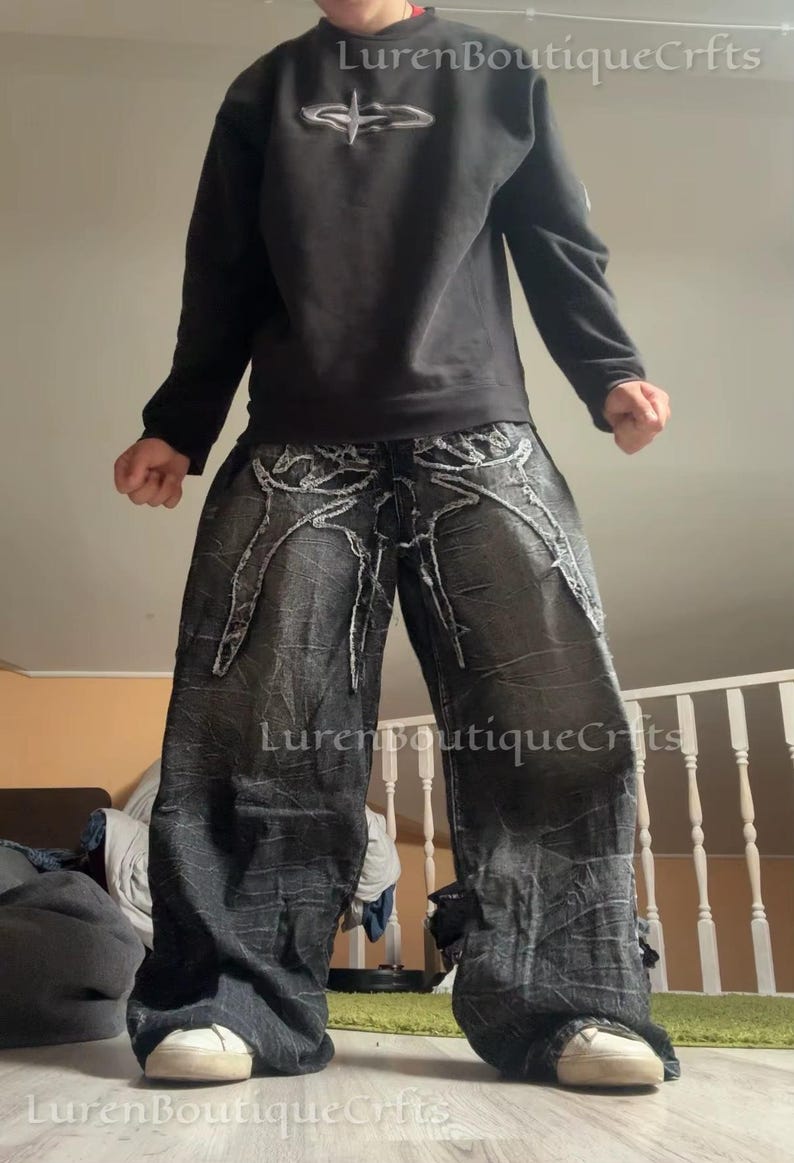 May include: Black sweatshirt with a silver emblem and distressed black denim jeans. The jeans have white stitching and a wide leg design. White trainers complete the outfit. The text "LurenBoutiqueCrfts" is visible.