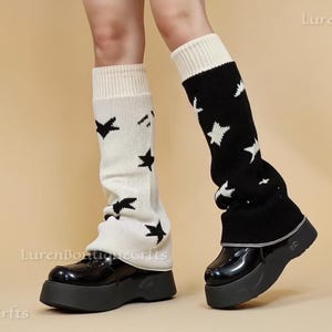 May include: A pair of platform shoes with black patent leather uppers. The legs are adorned with knee-high leg warmers, one cream-colored with black star patterns and the other black with white stars. The background is a solid tan color.