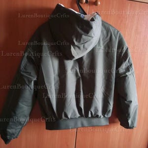 May include: Olive green hooded jacket with a white interior lining. The jacket has a ribbed hem and cuffs, and is hanging on a black hanger. The jacket is made of a smooth, possibly water-resistant material.