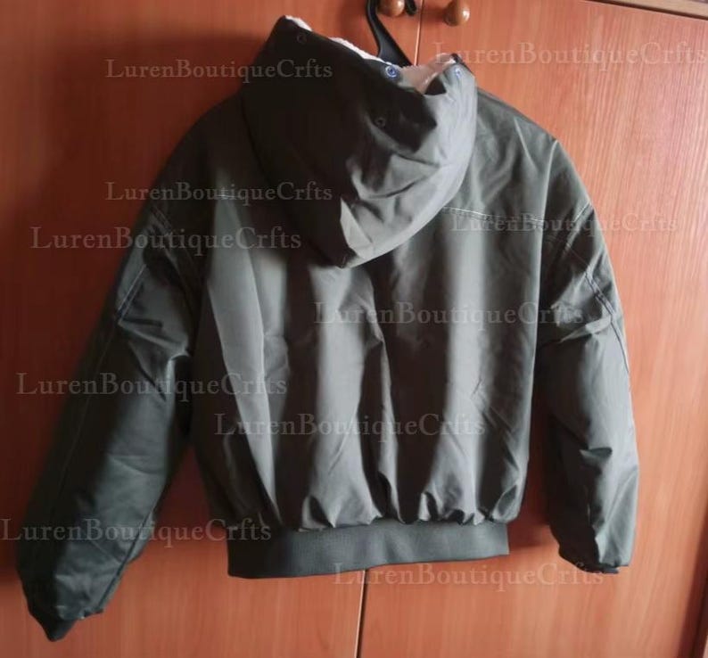 May include: Olive green hooded jacket with a white interior lining. The jacket has a ribbed hem and cuffs, and is hanging on a black hanger. The jacket is made of a smooth, possibly water-resistant material.