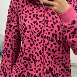 Puede incluir: Una sudadera con capucha rosa con cremallera y estampado de leopardo negro. La sudadera tiene una capucha forrada de piel sint&eacute;tica negra, una cremallera frontal y dos bolsillos delanteros. Los pu&ntilde;os y el dobladillo son de color rosa liso.