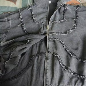 Puede incluir: Chaqueta con capucha de mezclilla gris con cierre de cremallera. La chaqueta presenta bordes deshilachados decorativos a lo largo de las costuras y alrededor de la capucha. La chaqueta es de mezclilla suave.