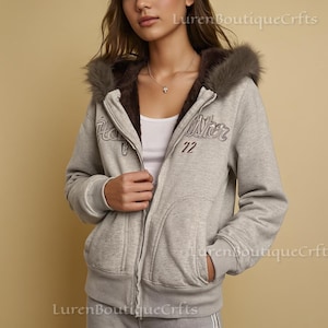 May include: A light grey zip-up hoodie with a faux fur-lined hood and a brown interior. The hoodie features the word "Hollister" and the number "72" embroidered on the front. The model is wearing a white vest underneath.