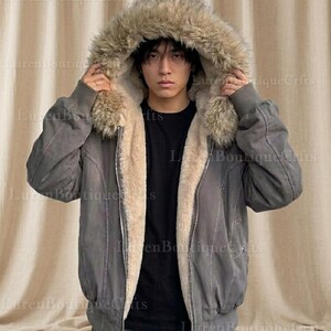 May include: A gray bomber jacket with a faux fur-lined hood. The jacket has a light beige interior lining and a zipper closure. The model is wearing a black shirt underneath. The jacket is a stylish outerwear option.